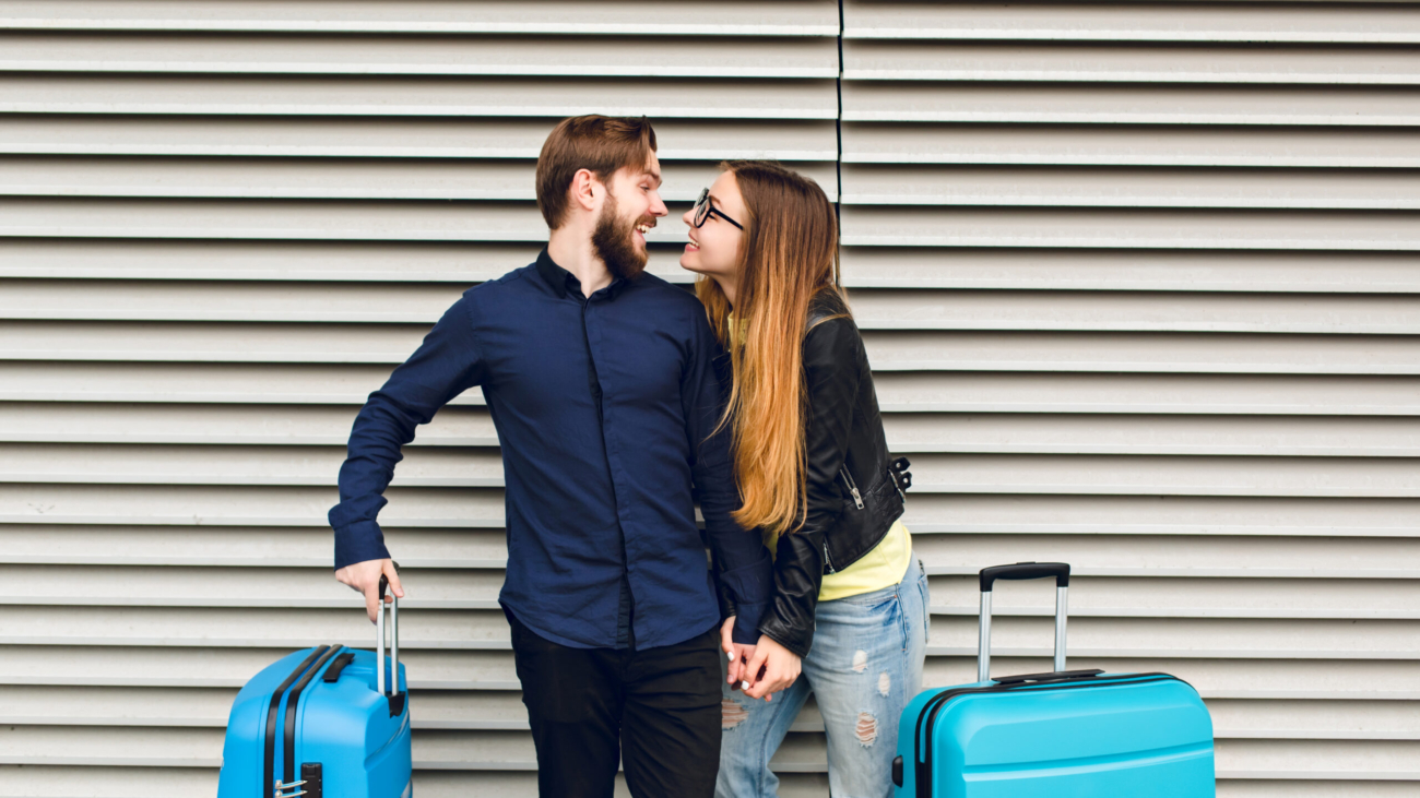 Portrait of cute couple  standing on gray  striped background.  She has long hair, glasses,  yellow sweater, jacket. He wears black shirt, beard. They are  hugging and smilinhg to each other.