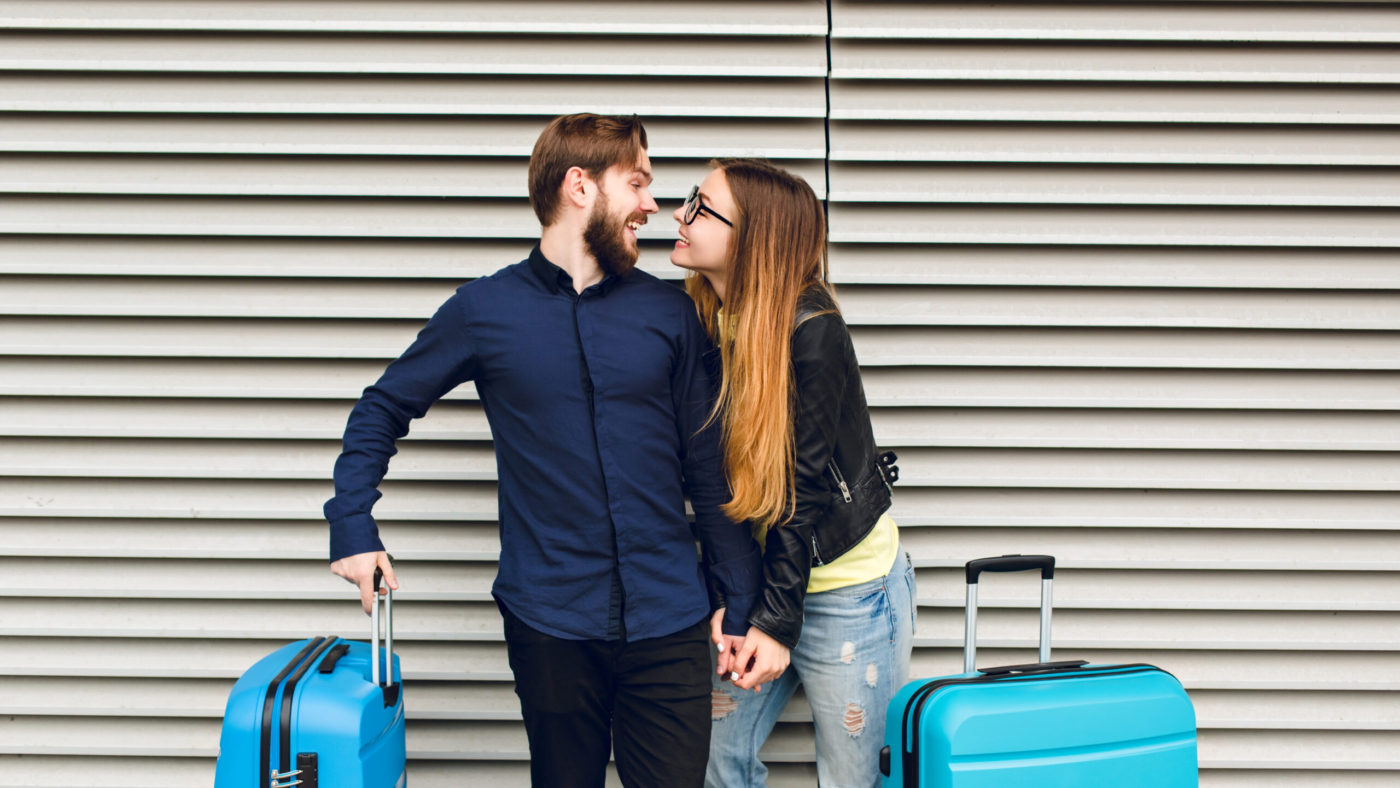 Portrait of cute couple standing on gray striped background. She has long hair, glasses, yellow sweater, jacket. He wears black shirt, beard. They are hugging and smilinhg to each other.